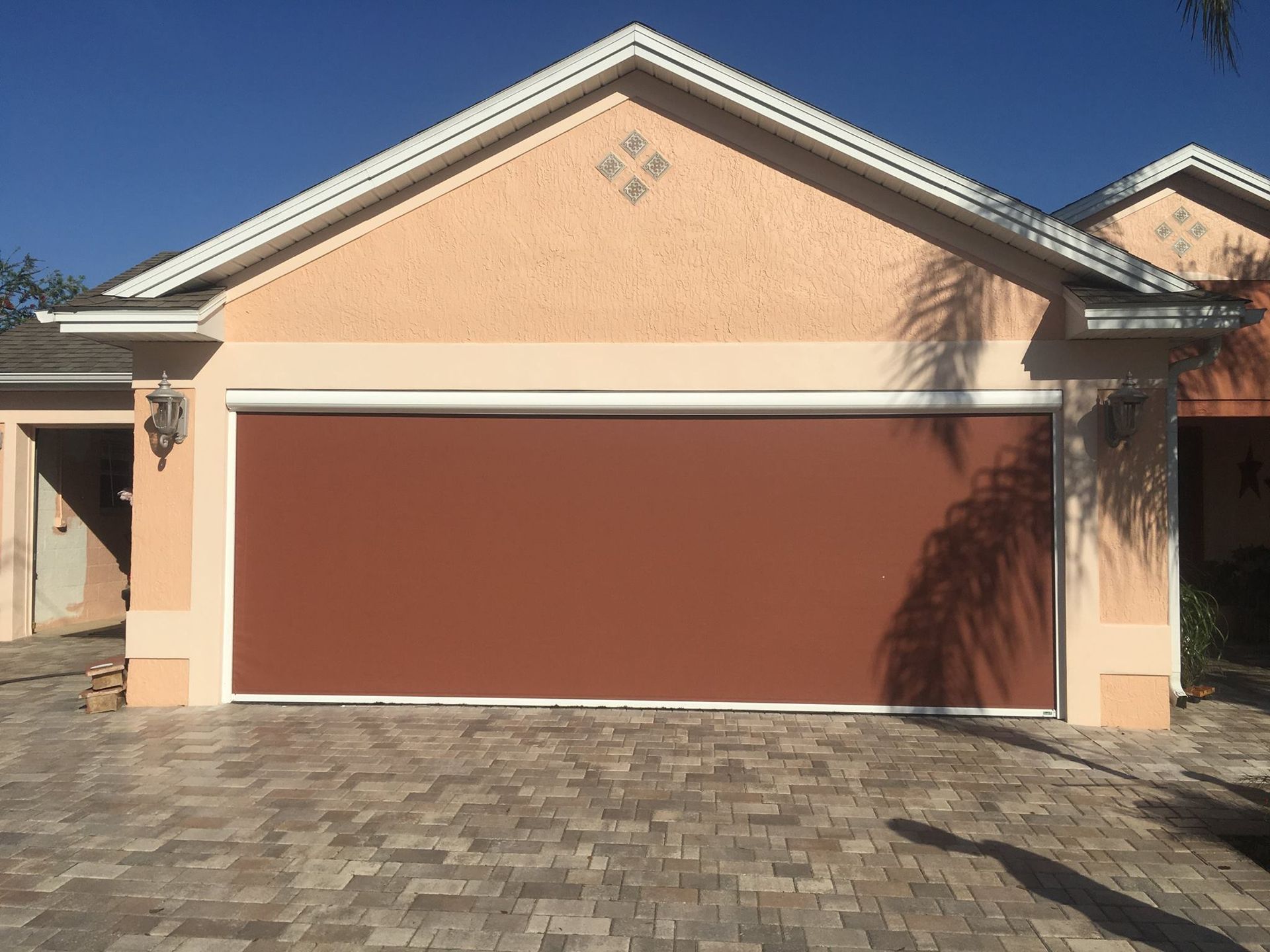 A beige house garage door covered by a solid, brown roll-down screen, situated on a stone-paved driveway.