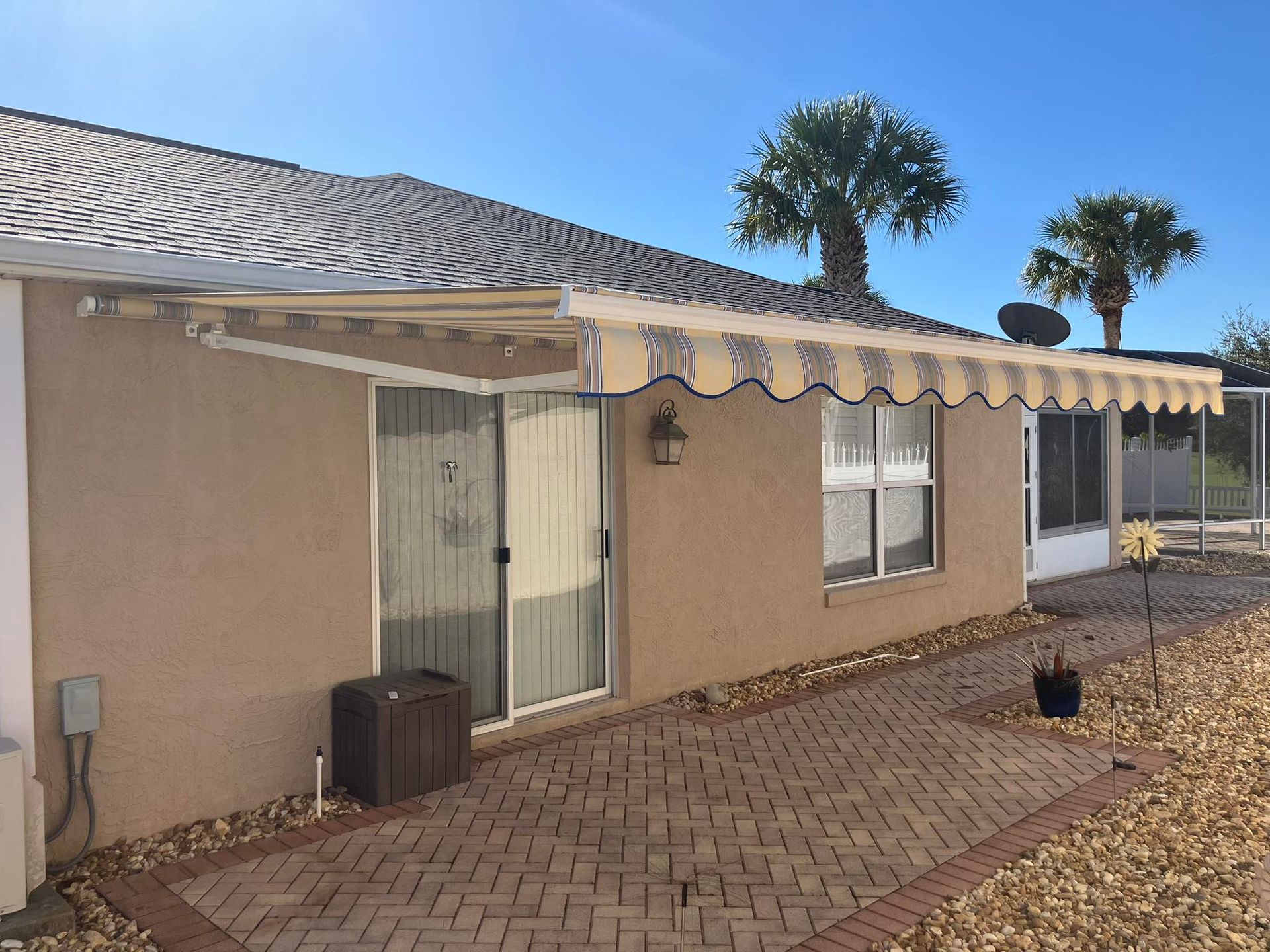 A tan stucco house with a retractable cream and brown striped awning over a glass sliding door and brick patio.