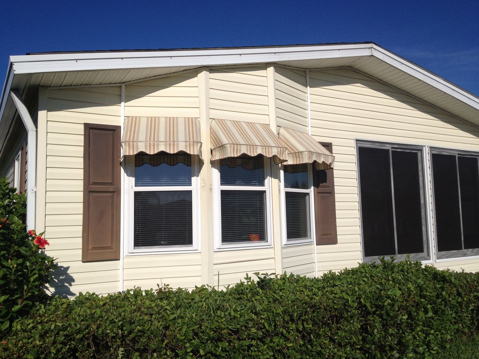 A beige mobile home exterior with striped window awnings, dark brown shutters, and green bushes in the foreground.