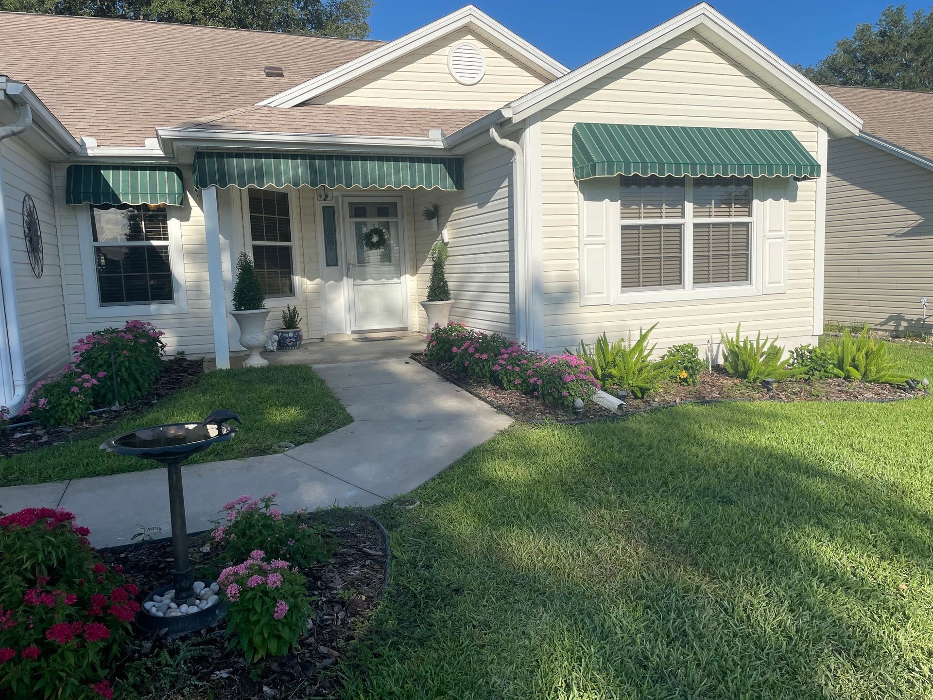 A single-story, beige house with a green roof awning, a paved walkway, and a landscaped yard on a sunny day.