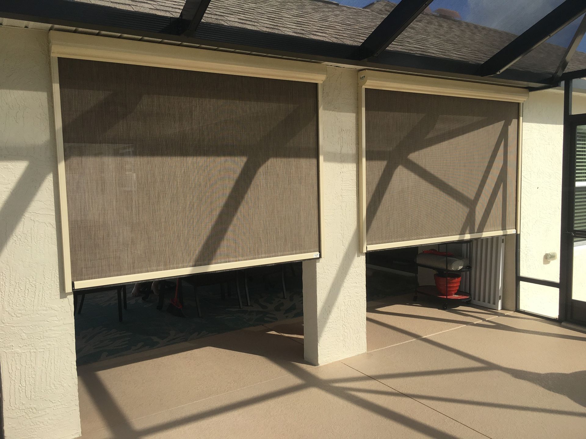 Two brown, retractable solar shades installed on a beige stucco patio enclosure under a screened roof.