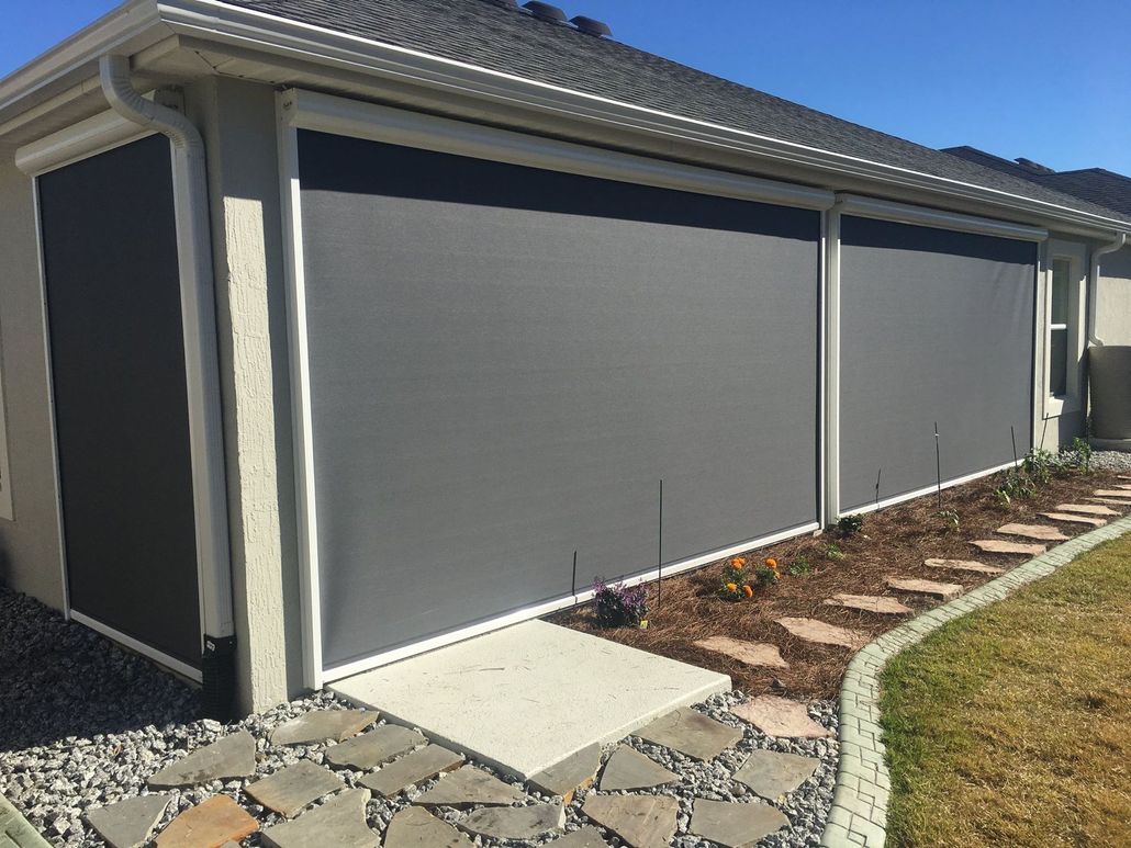 Exterior view of a beige house featuring retractable dark gray solar screens covering the patio and side wall.