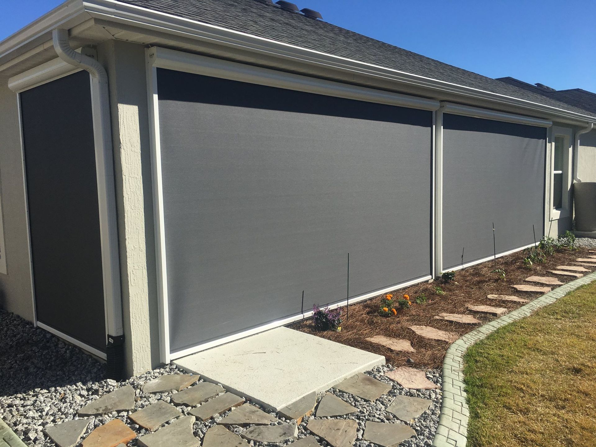 Exterior view of a beige house featuring retractable dark gray solar screens covering the patio and side wall.