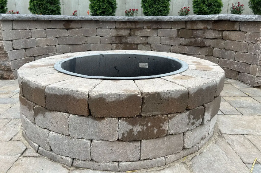 Circular brick fire pit with metal insert, on a paved patio, with a brick wall behind it.