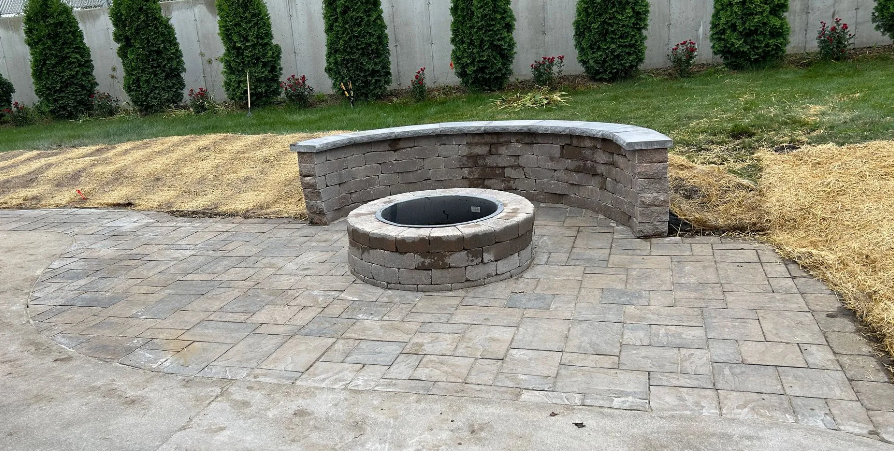 Brick patio with a built-in fire pit and a low curved wall in a backyard with green grass and trees.