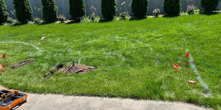Lawn with white lines marking an area for digging, with green grass, small trees in the background, and red flags.