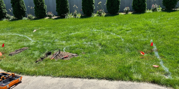 Lawn with white lines marking an area for digging, with green grass, small trees in the background, and red flags.