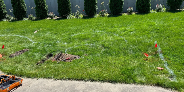 Lawn with white lines marking an area for digging, with green grass, small trees in the background, and red flags.