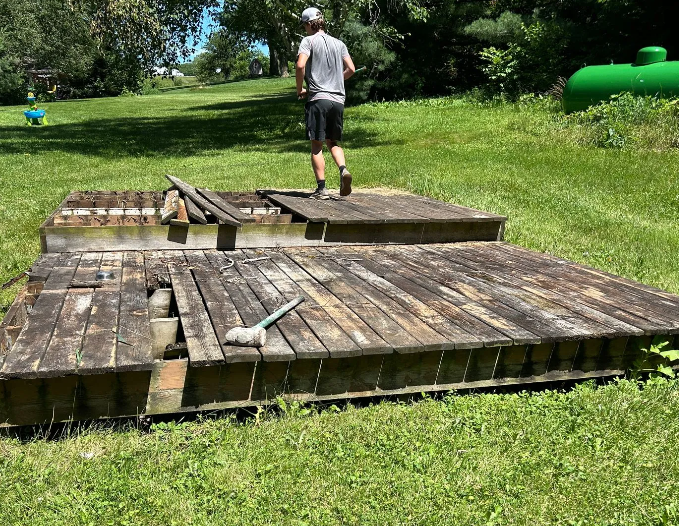 Man walks across a weathered wooden dock in a grassy yard, with a green propane tank visible.