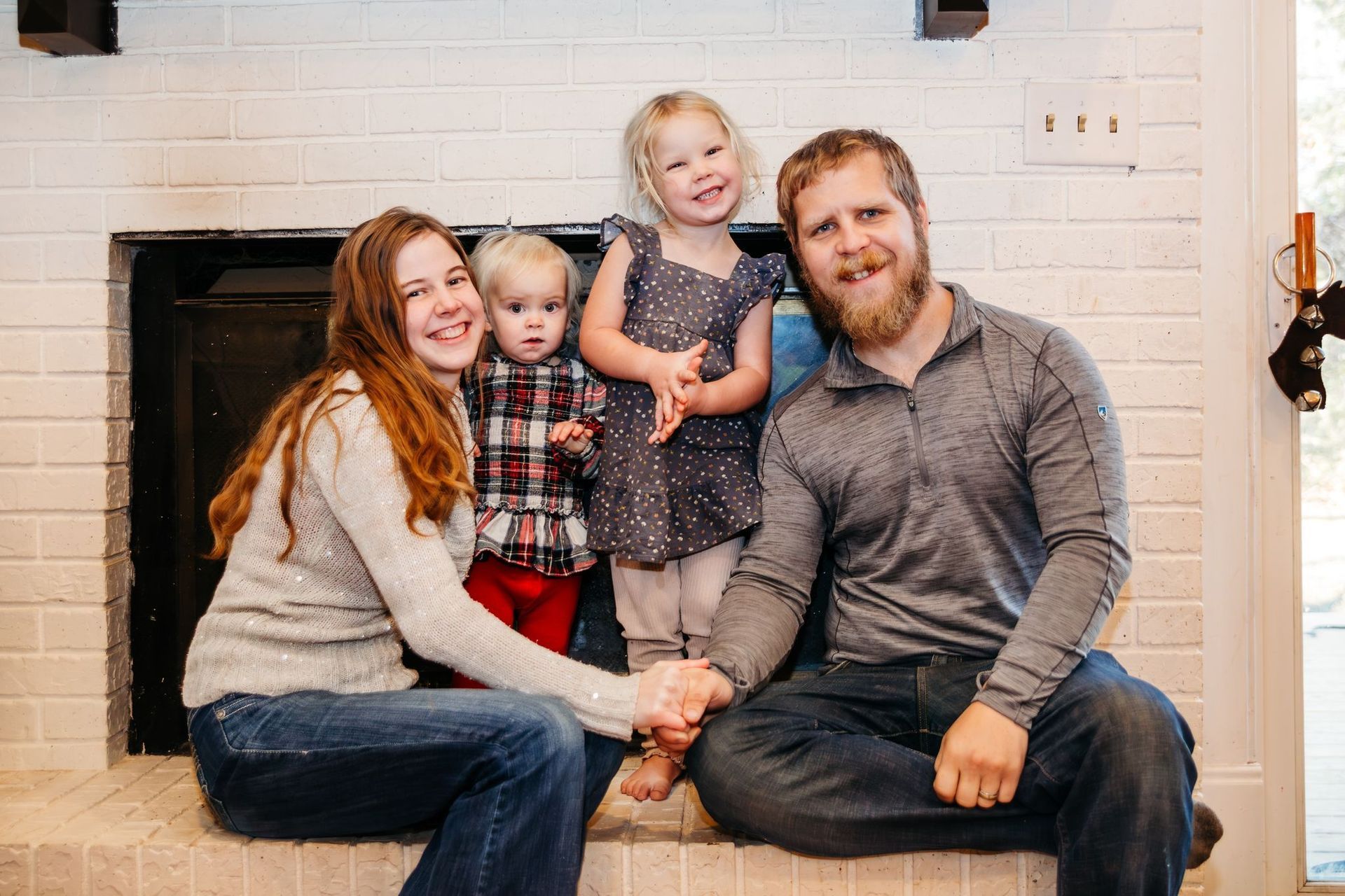 Family of four smiling in front of a white brick fireplace.
