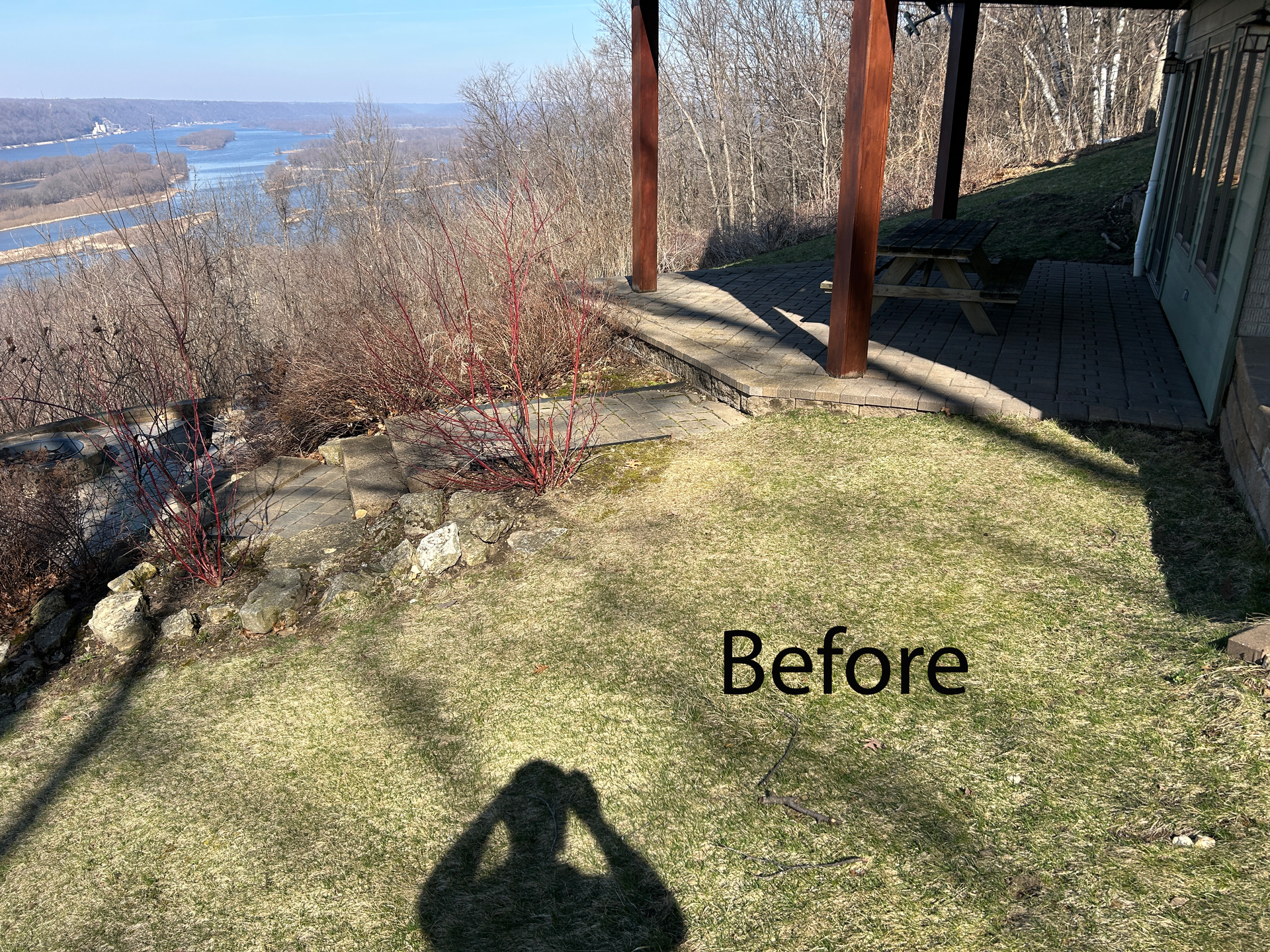 Grassy yard beside river with bare shrubs, wooden posts, and a shaded patio; “Before” text overlay.