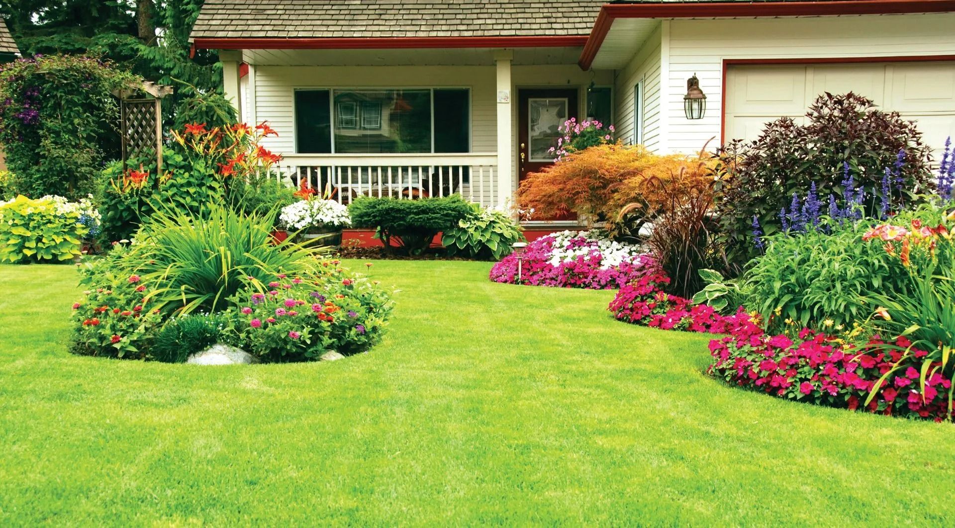 Green lawn with colorful flower beds in front of a white house with a garage and porch.