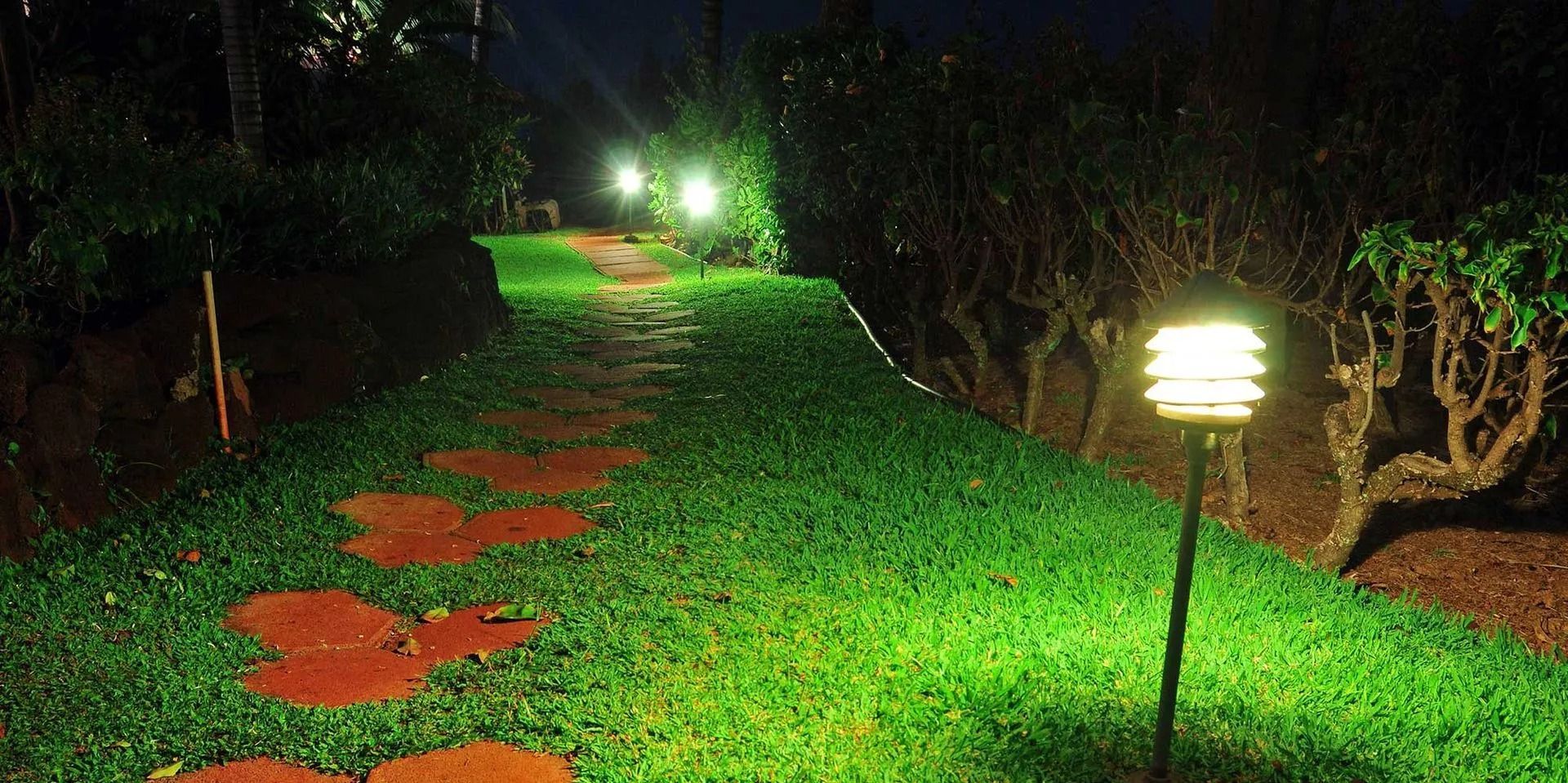 A stone path illuminated by lights leads through a grassy garden at night.