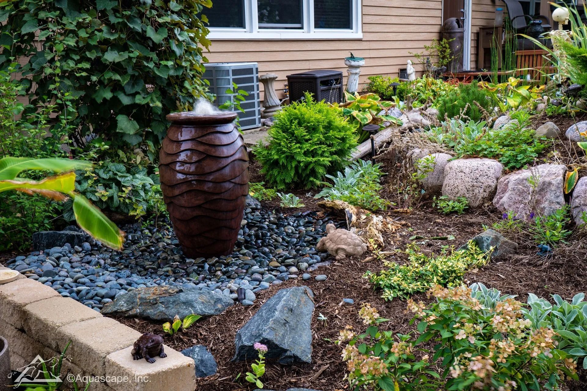Water fountain in a landscaped garden with rocks, plants, and a house in the background.