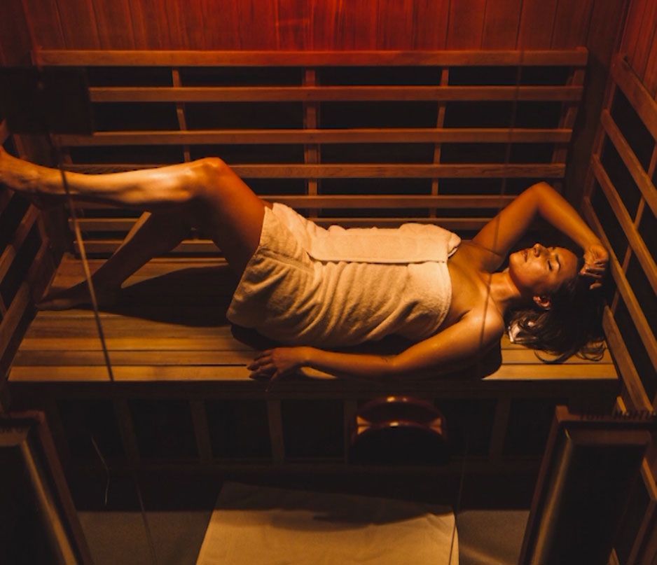 Woman relaxes in a sauna, wrapped in a towel, lying on wooden benches in dim red light.