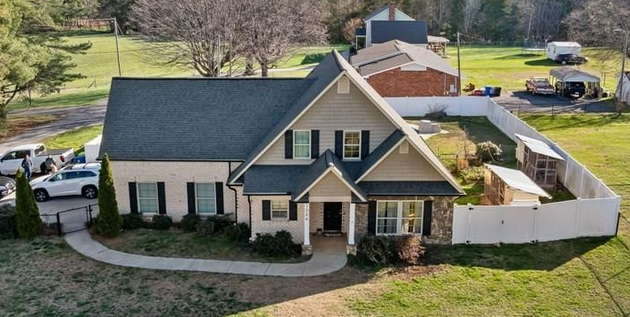 An aerial view of a suburban home with tan siding, a dark roof, and a white fenced backyard on a sunny day.