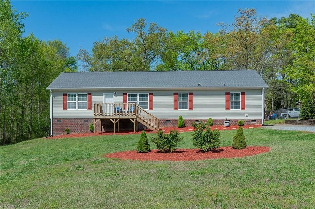 A one-story ranch-style house with light siding and brick foundation, featuring a front deck, set in a grassy yard.