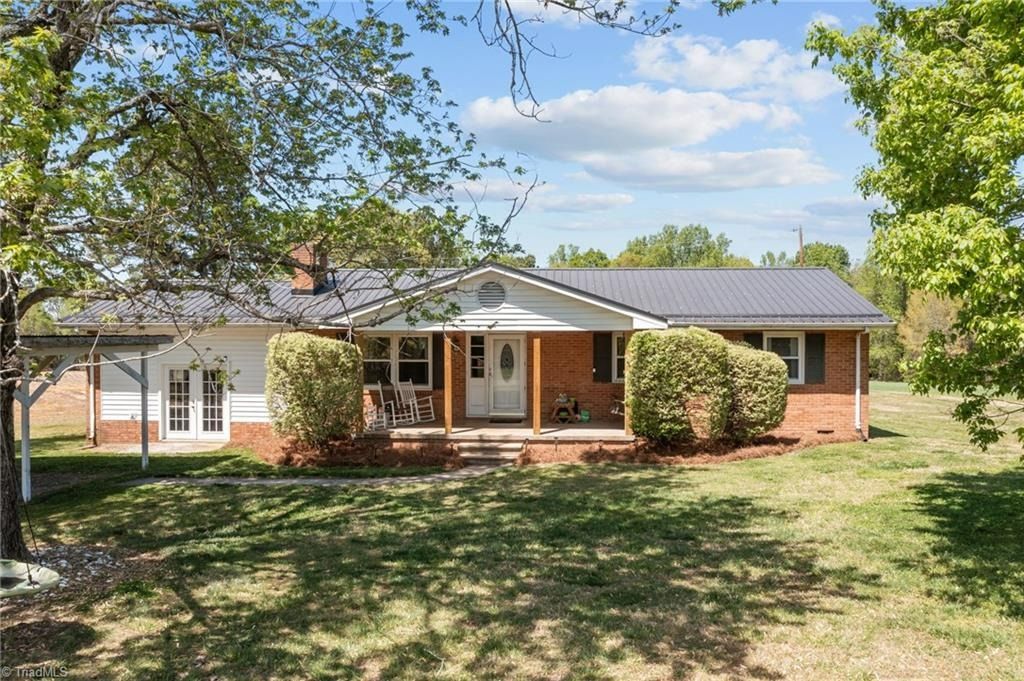A single-story ranch-style home with a brick exterior, a dark metal roof, and a front porch, surrounded by green trees.