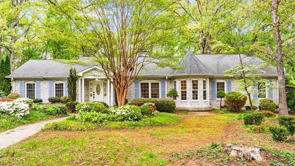 A single-story, light-colored brick house with blue shutters, surrounded by mature trees and lush green landscaping.