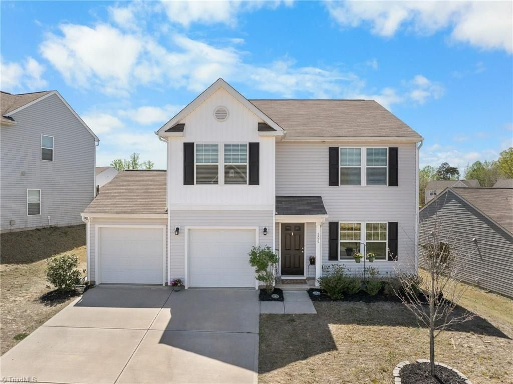 A two-story suburban home with light gray siding, white trim, a two-car garage, and a brown shingle roof under a blue sky.