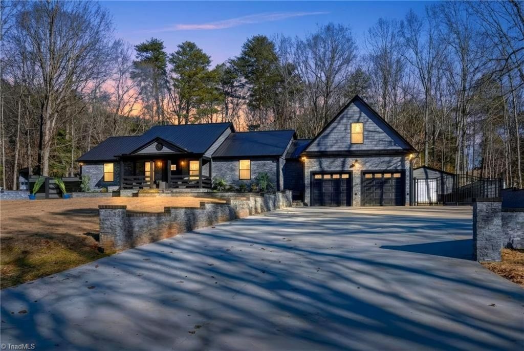 A dark-toned, single-story house with a two-car garage and stone facade, nestled in a wooded area at dusk.