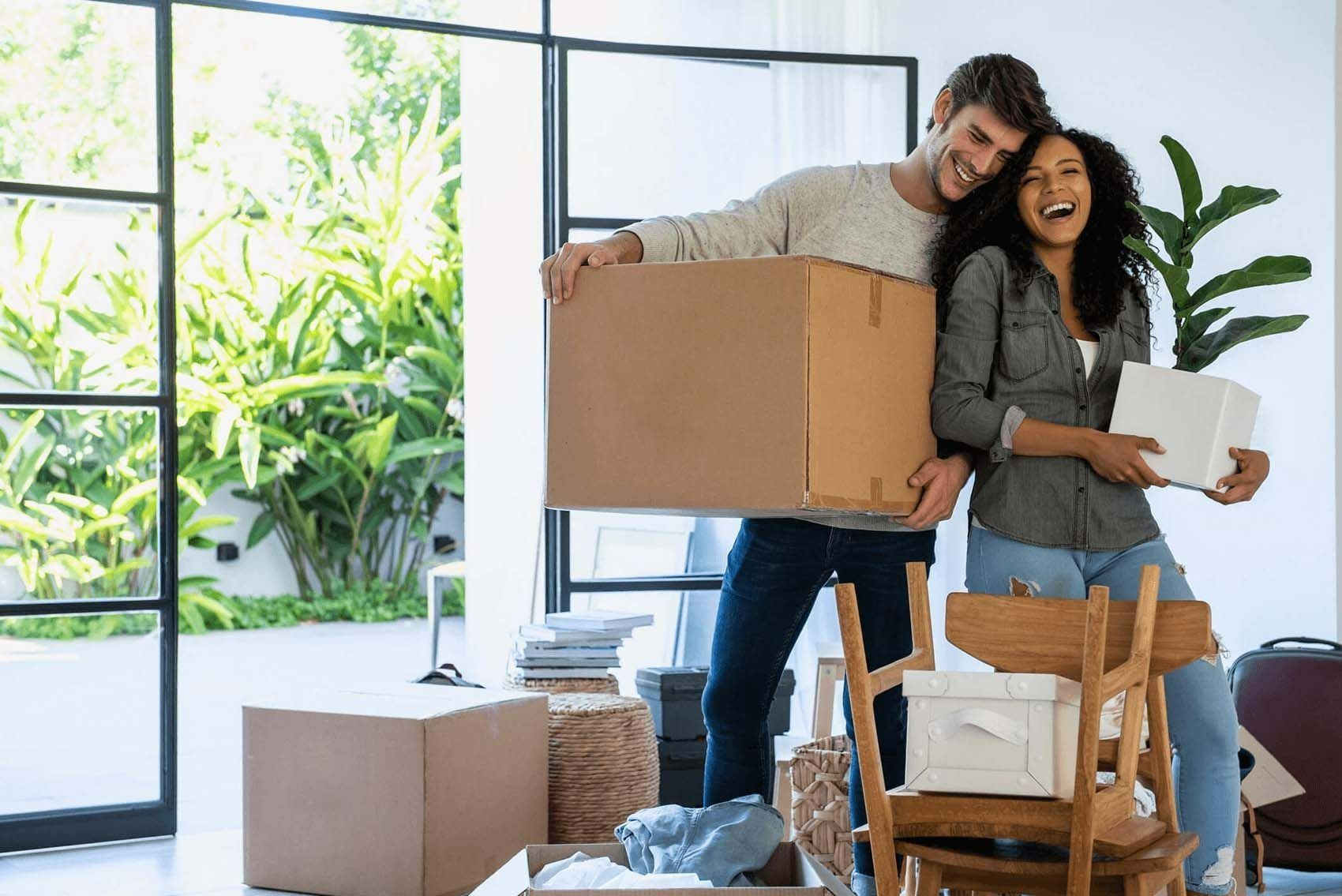 A smiling couple holding boxes and a plant in a bright, modern room filled with cardboard boxes, signaling a move.