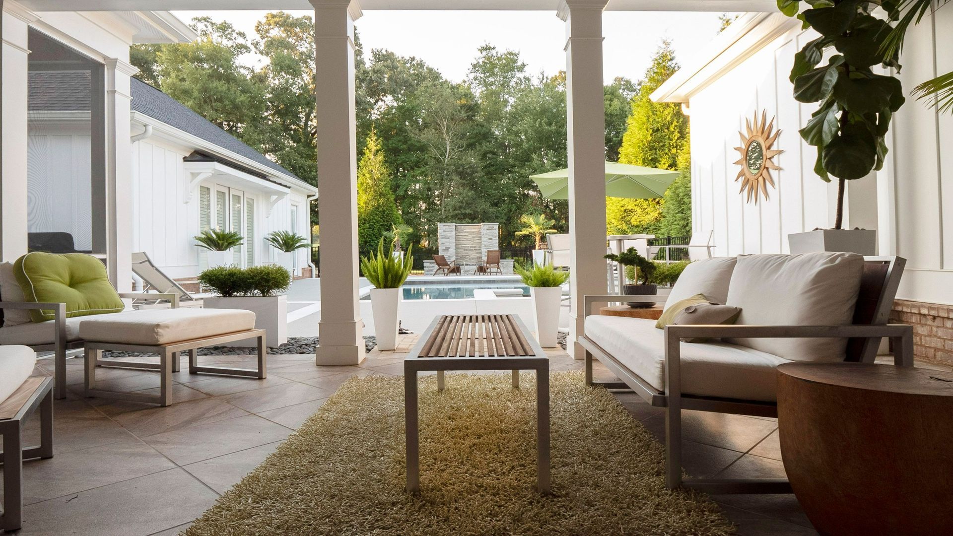 A covered outdoor patio featuring white furniture, a textured rug, and a view of a backyard pool and lush greenery.