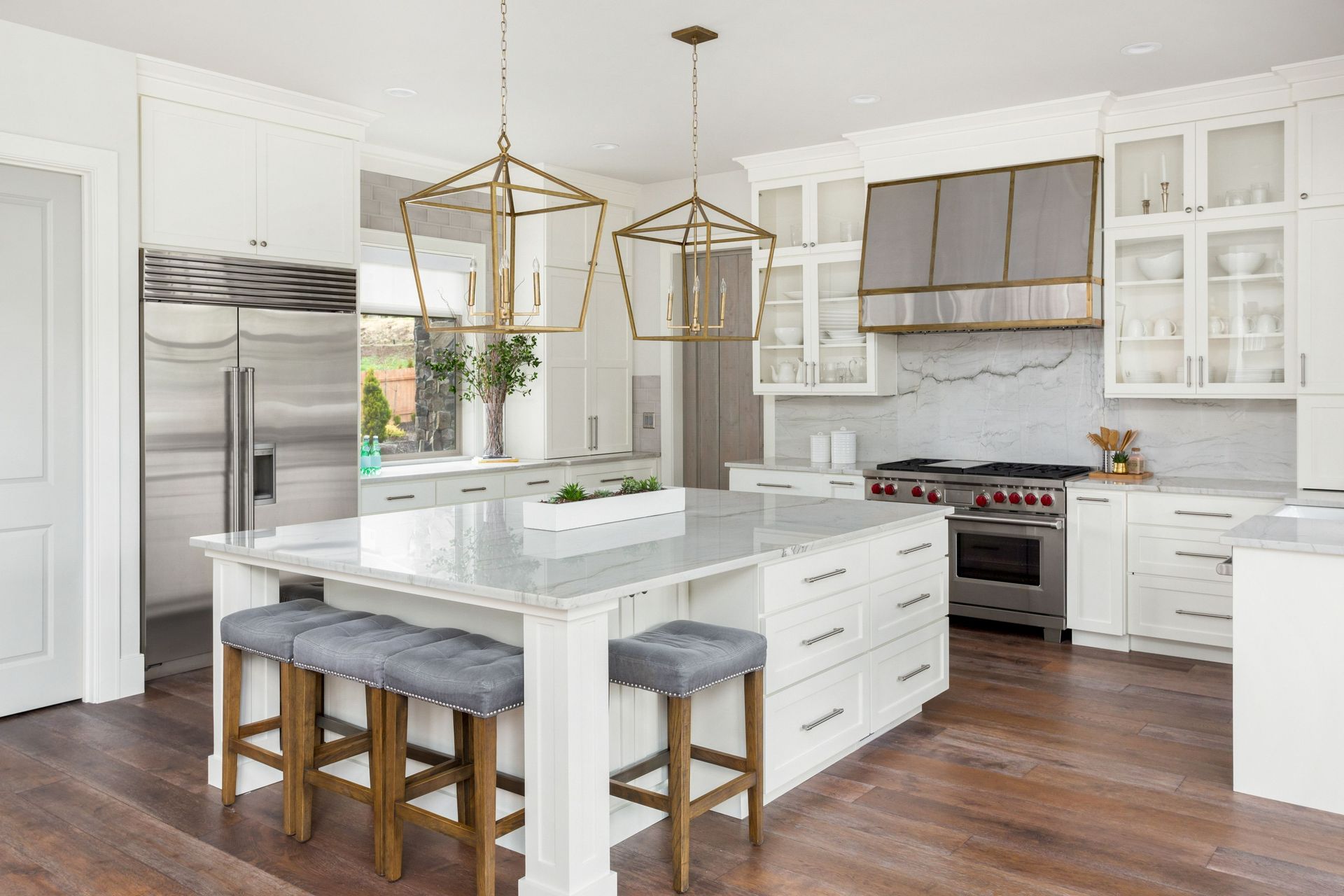 A modern white kitchen featuring a large central island, gray bar stools, stainless steel appliances, and wood flooring.
