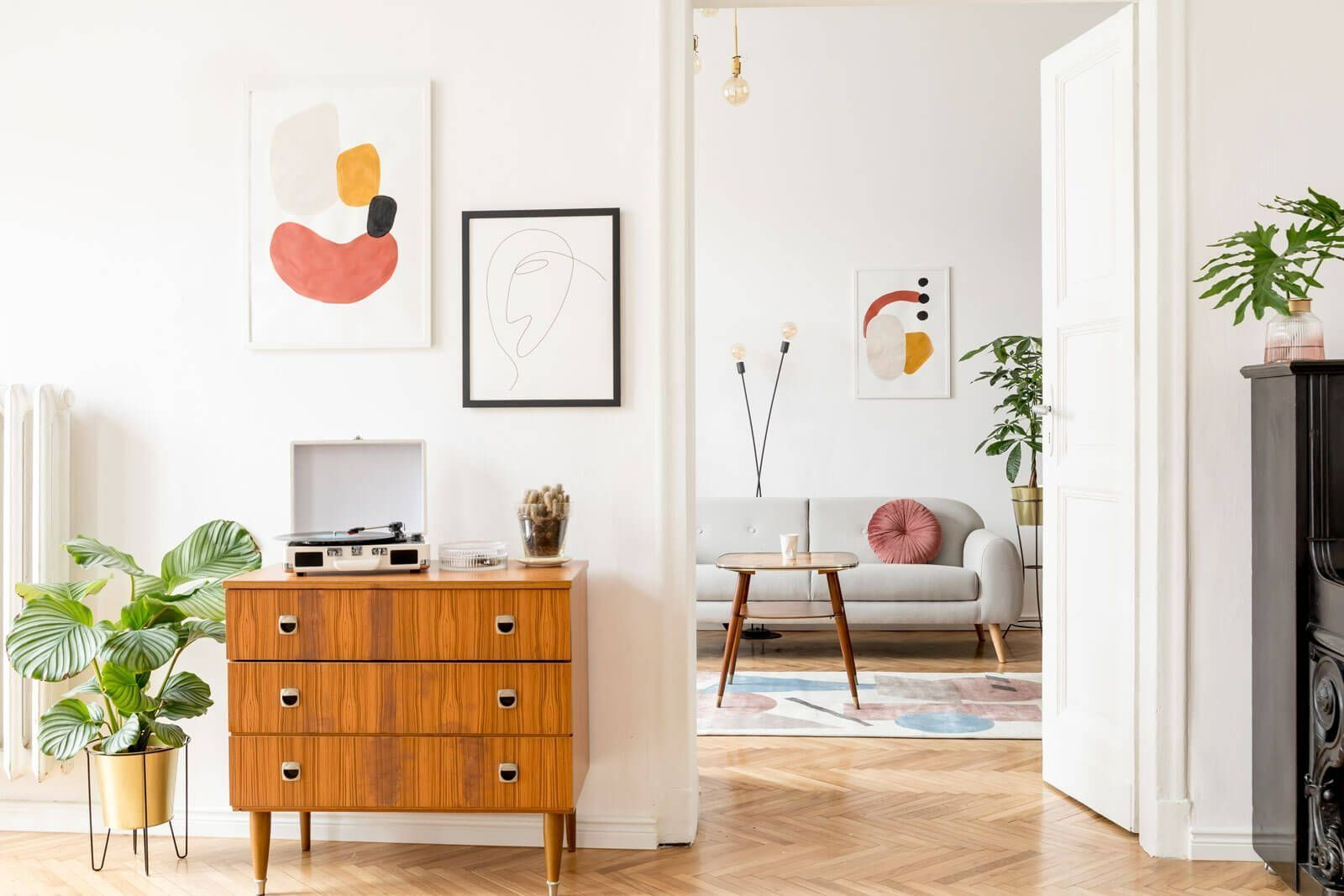 A wooden mid-century dresser with a record player sits in a white room, viewing into a living room with a gray sofa.