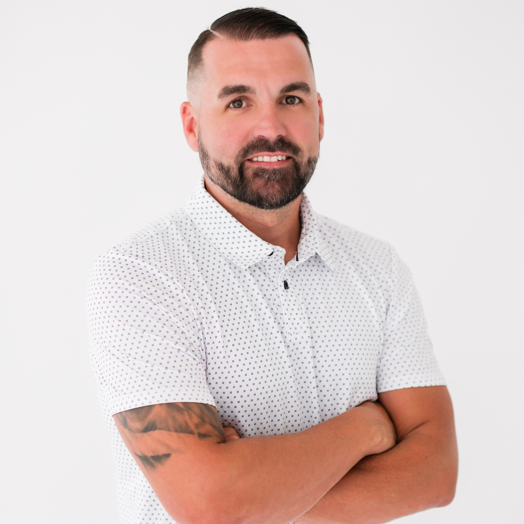 A person with a beard and a tattooed arm, smiling with arms crossed, wearing a white patterned polo against a white wall.