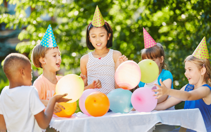 Children at a birthday party, holding balloons, wearing party hats outdoors.