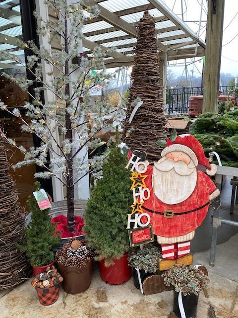 Holiday garden display with Santa cutout, potted trees, and festive decorations under a greenhouse canopy