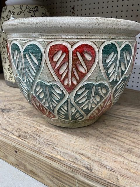 Ceramic bowl with carved leaf and heart design, painted red and teal, on a wooden shelf.