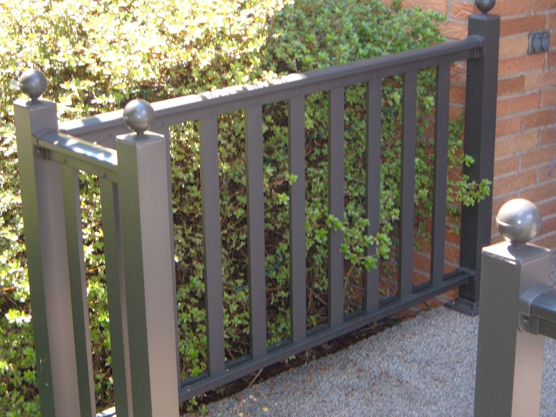 Black metal gate with vertical bars beside a brick wall and greenery behind it