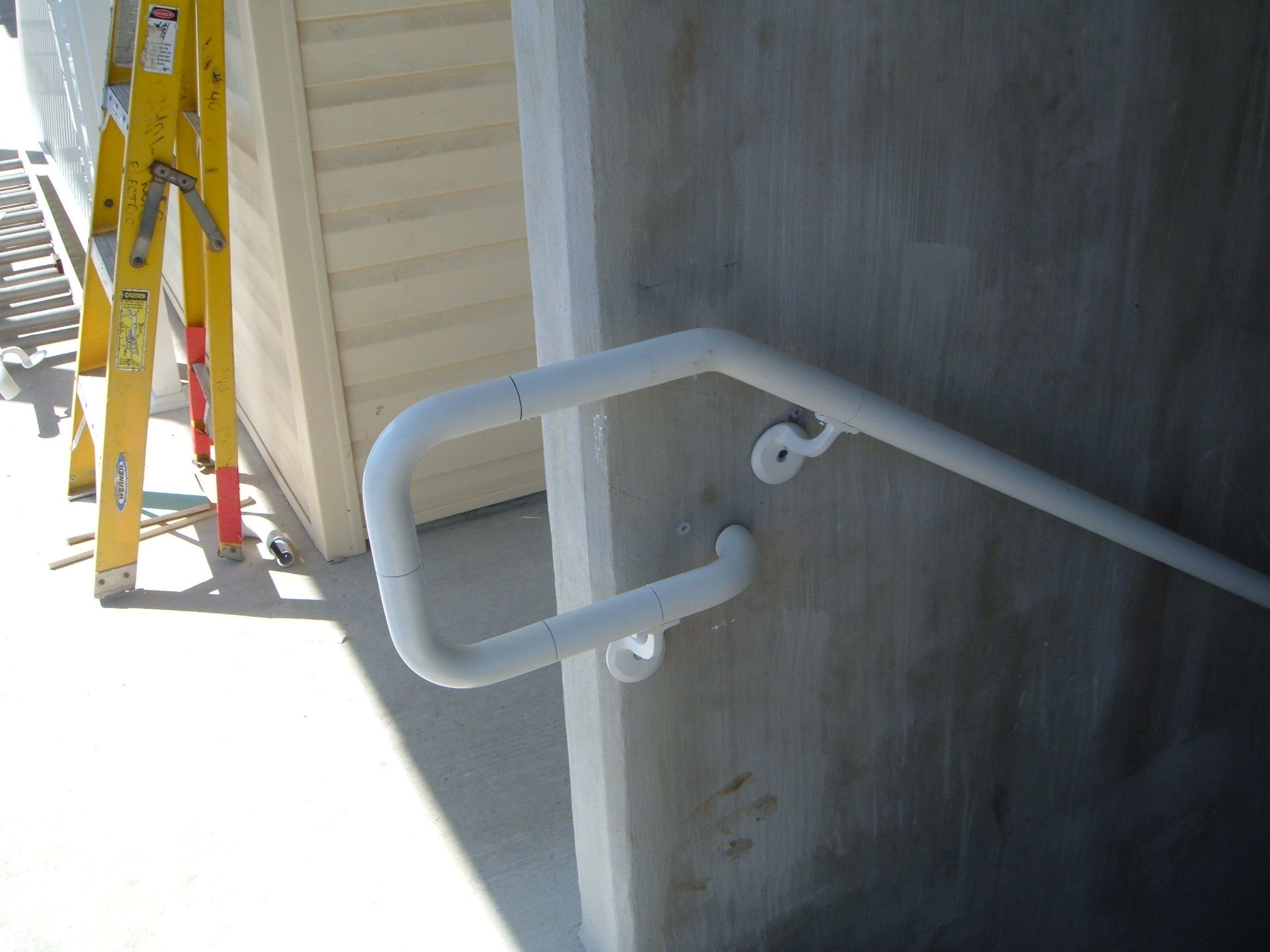 White handrail beside a concrete wall next to yellow ladders on a bright exterior walkway
