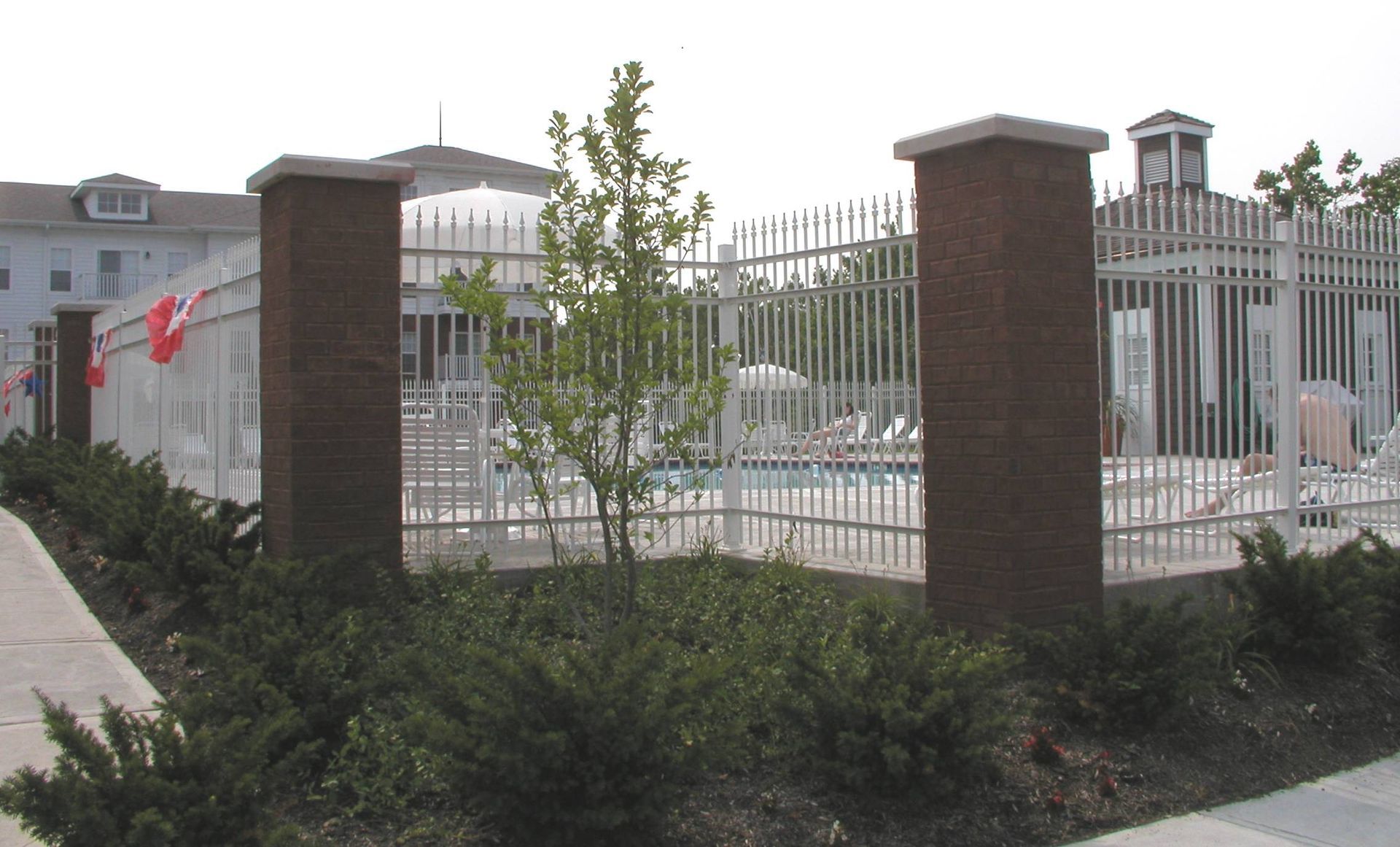Gated outdoor pool area with white fence, brick pillars, and a cloudy sky