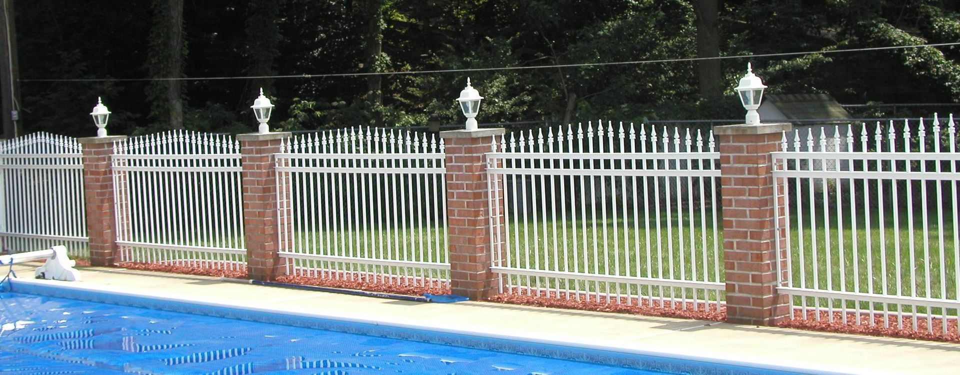 Brick-and-white metal fence beside a blue swimming pool with trees in the background