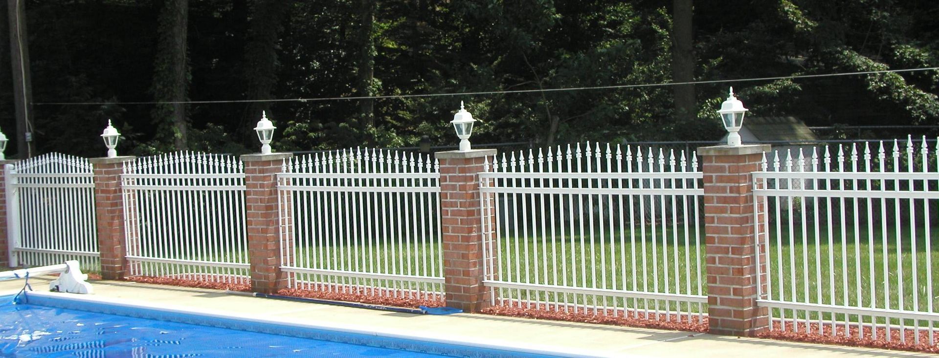 White metal fence with brick posts beside a blue pool in a landscaped yard