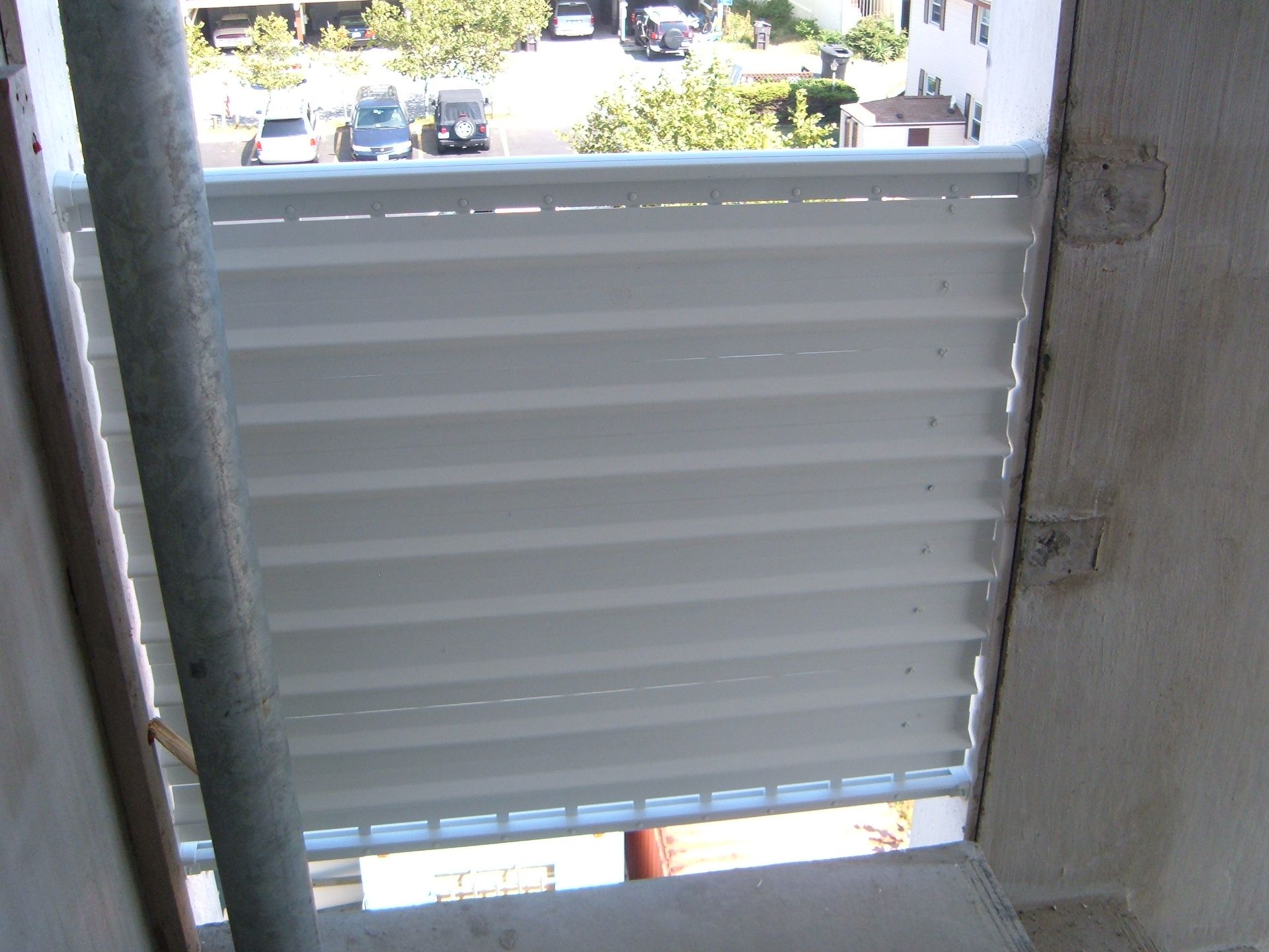 White louvered window shutter partially open, looking out to a bright street with parked cars.