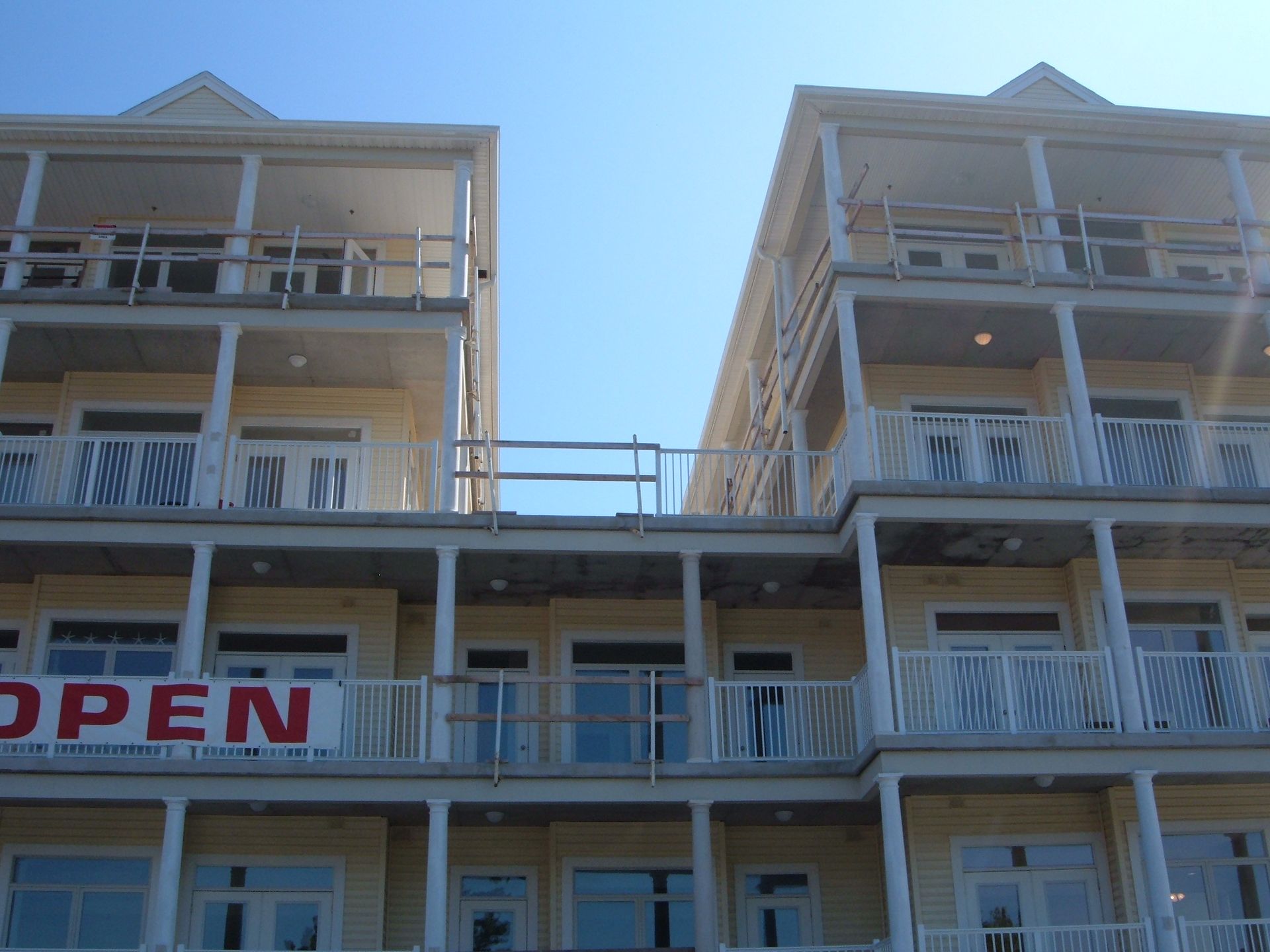 Two beige apartment balconies with white railings under a blue sky, with a red “OPEN” sign below