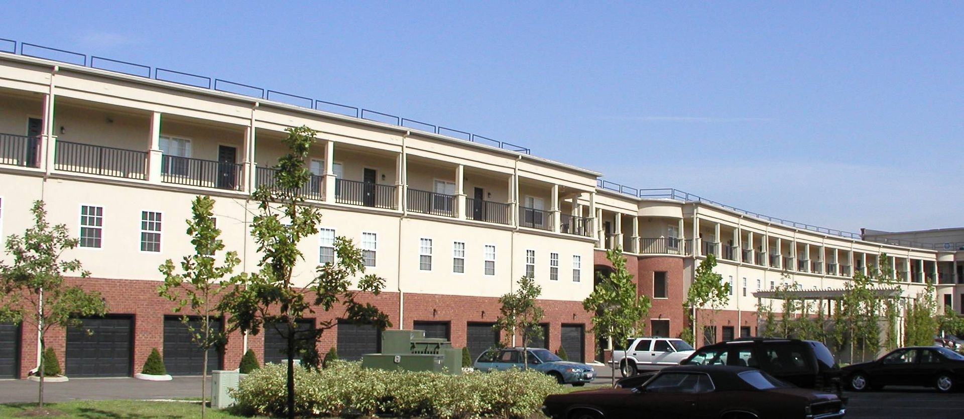 Modern building with white and brick facade, balconies, trees, and parked cars under a clear sky