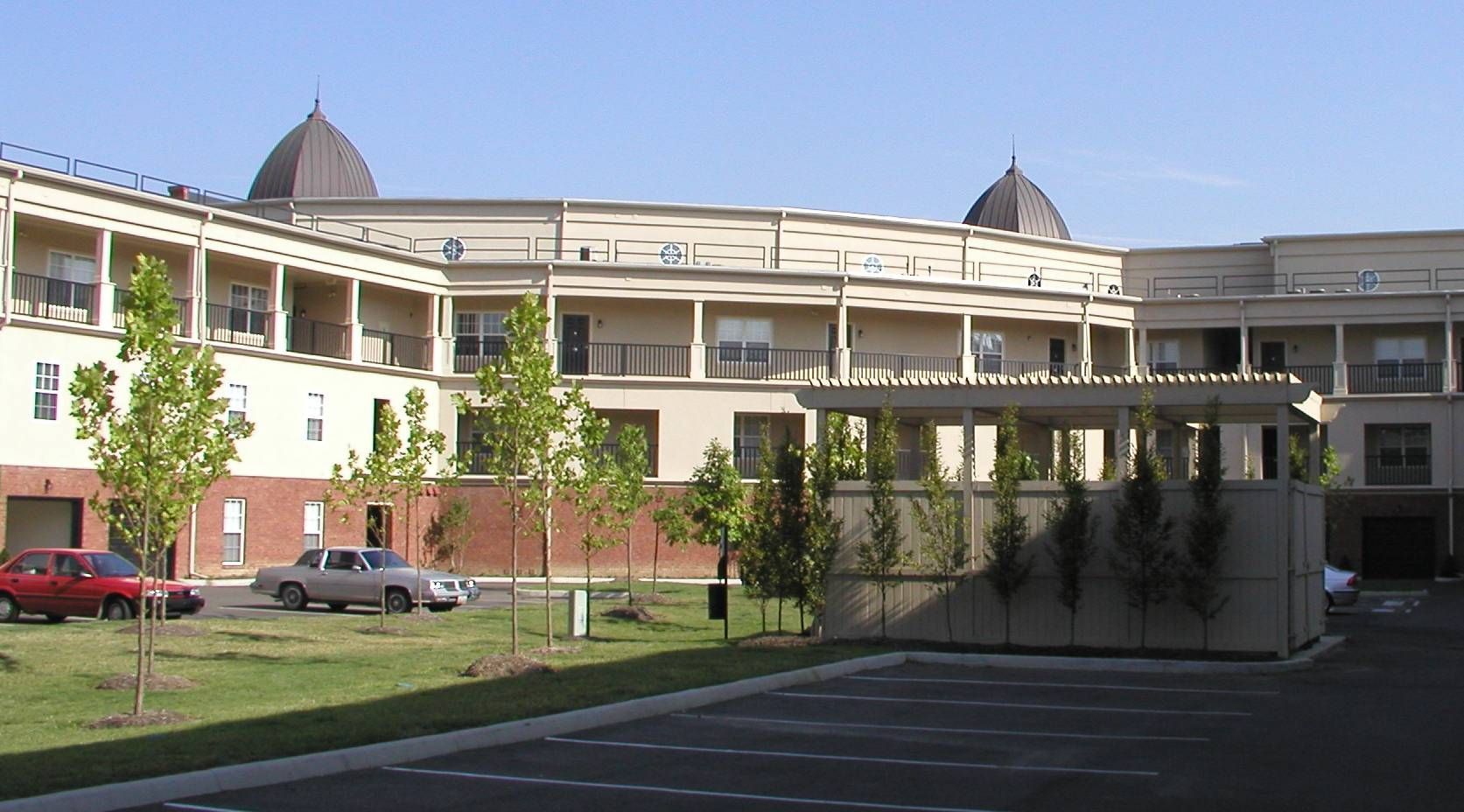 Apartment complex courtyard with white balconies, brick base, parked cars, and trees under a clear blue sky