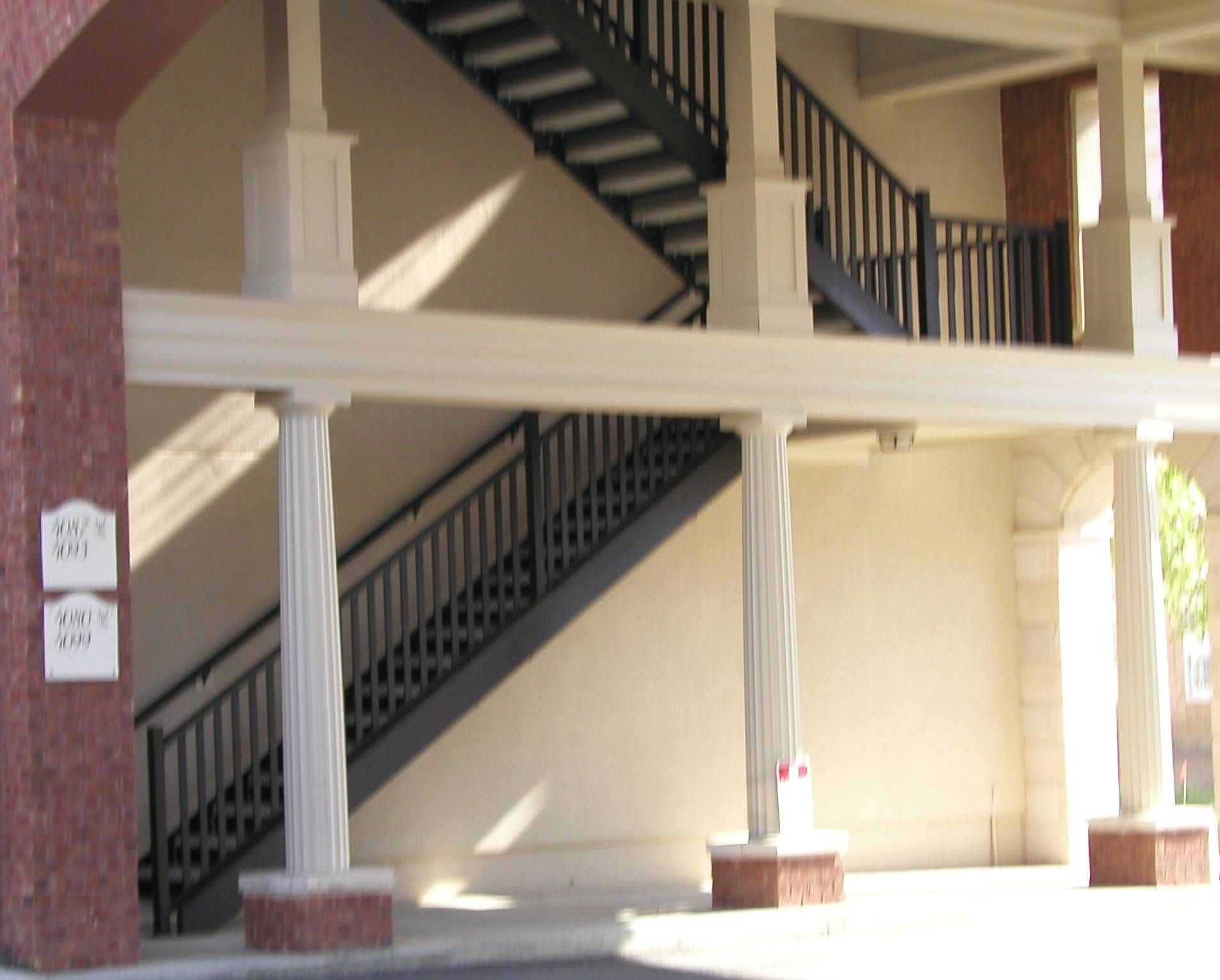 Covered outdoor stairway with black railings and beige columns on a brick walkway
