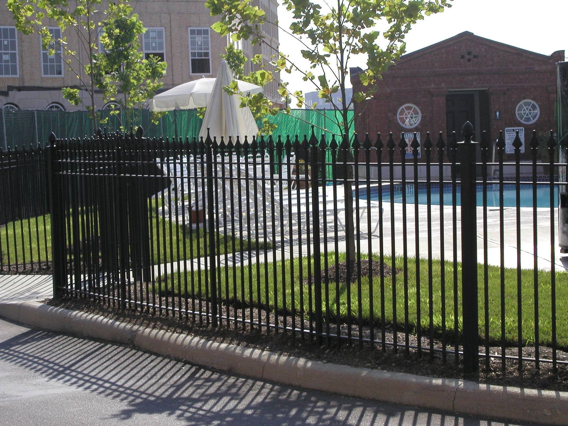 Black metal fence in front of a pool with umbrellas and a brick building in the background.