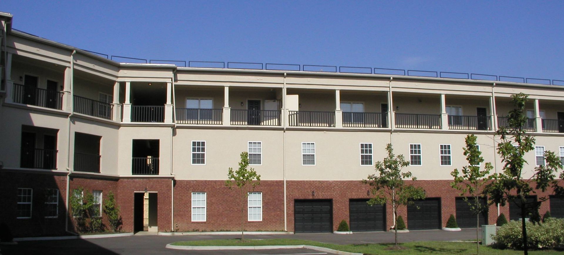 Three-story apartment building with white balconies, brick lower level, and trees in front under a blue sky