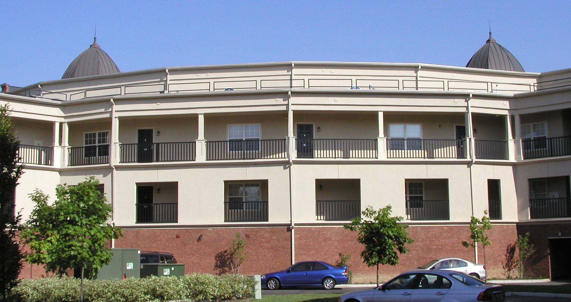 A curved cream and brick building with balconies, trees in front, and parked cars along the street.