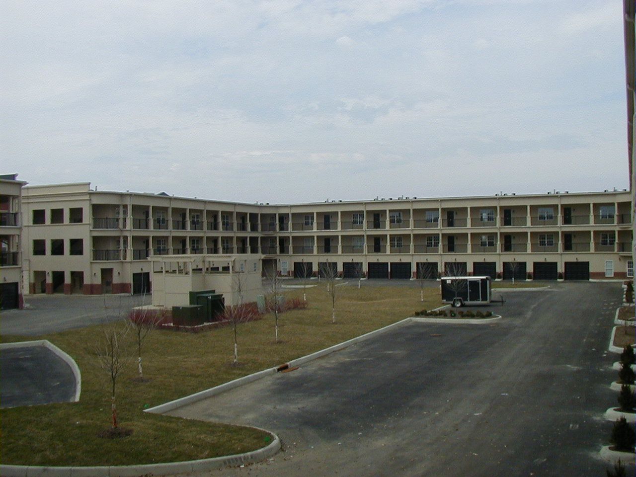 Large beige apartment building with balconies beside an empty paved lot and grassy courtyard under a cloudy sky