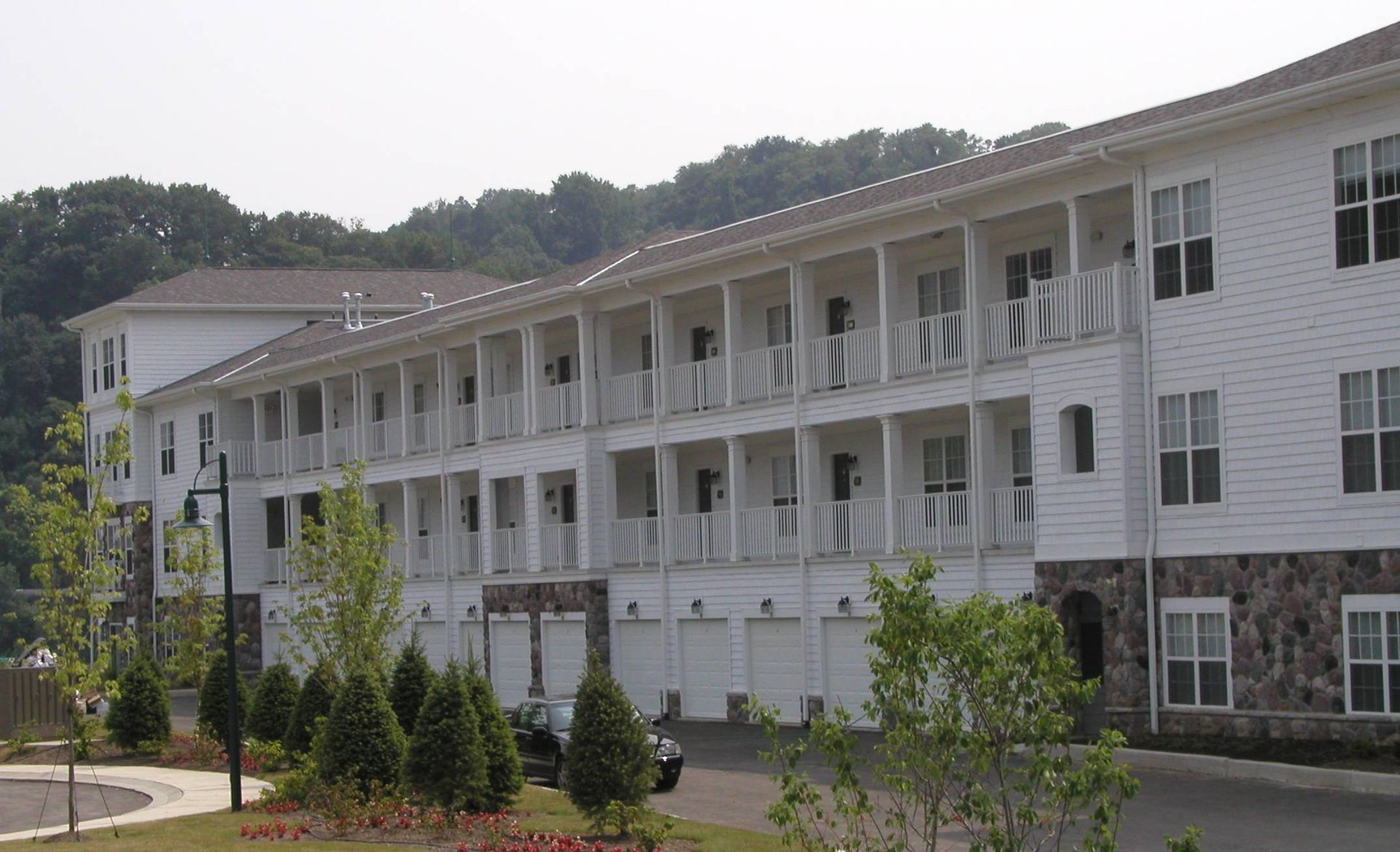 White multi-story apartment building with balconies and stone base beside a tree-lined road