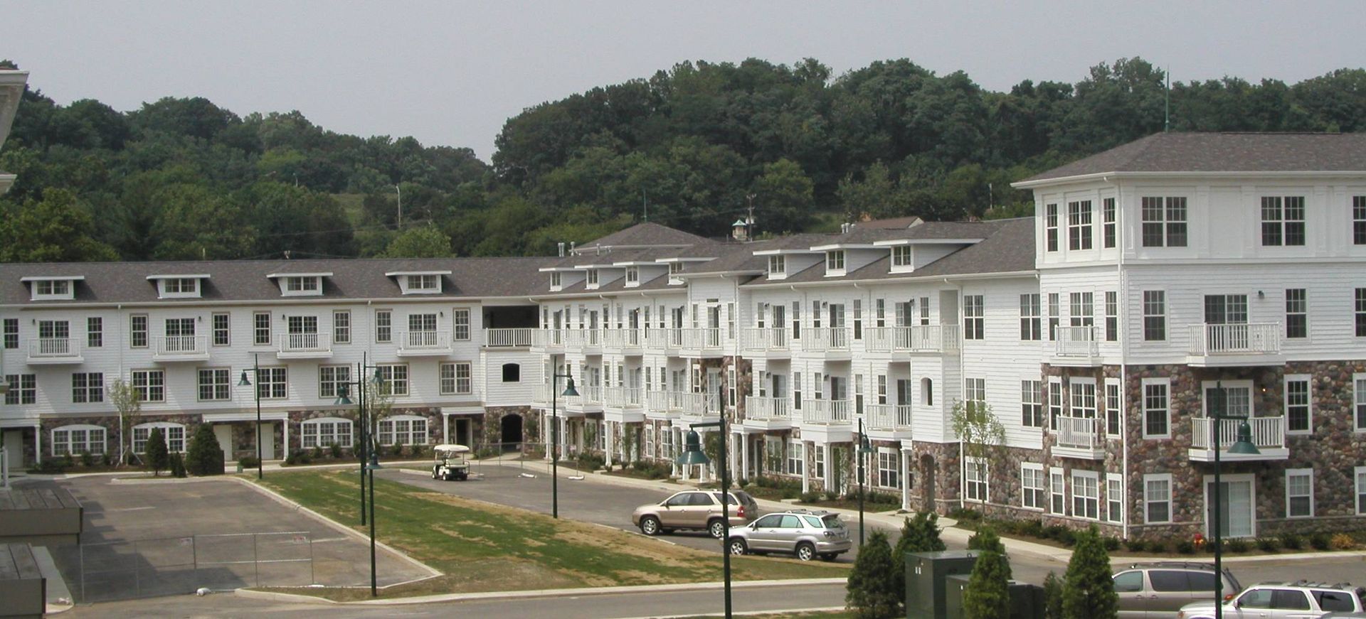Row of white townhouses with brick accents beside a paved lot and green hillside