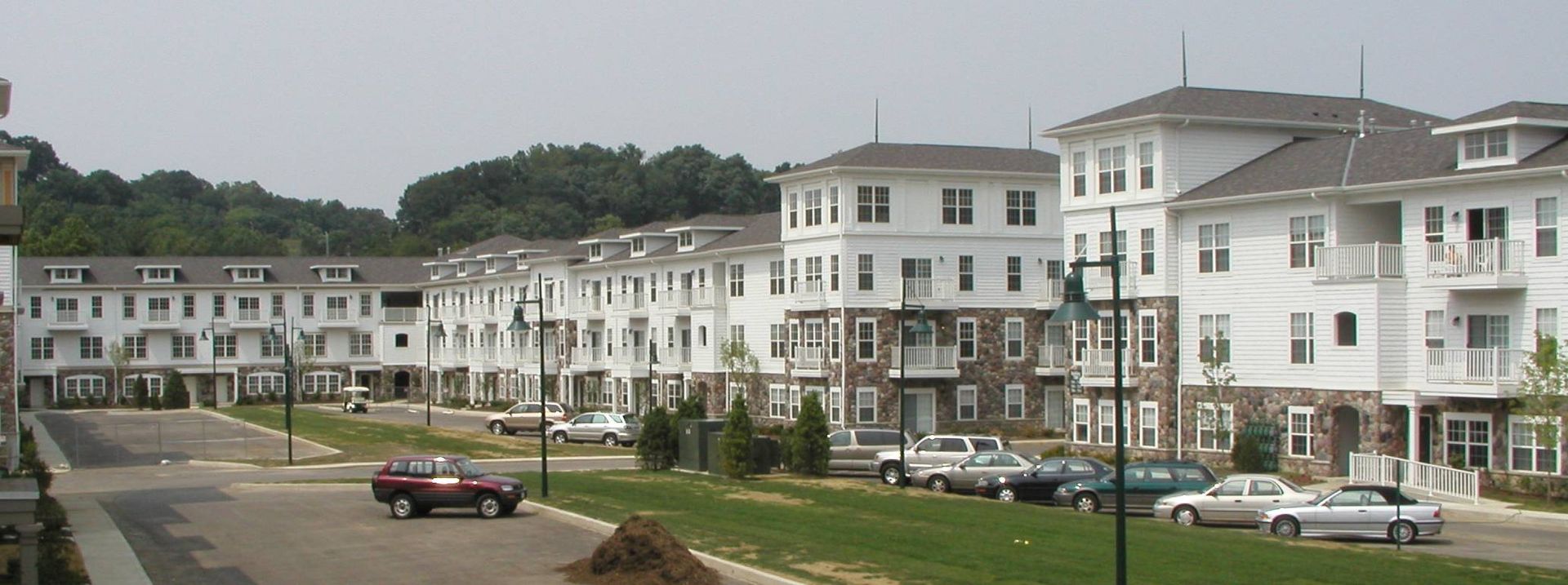 Row of white apartment buildings along a street with parked cars and grassy hills behind them